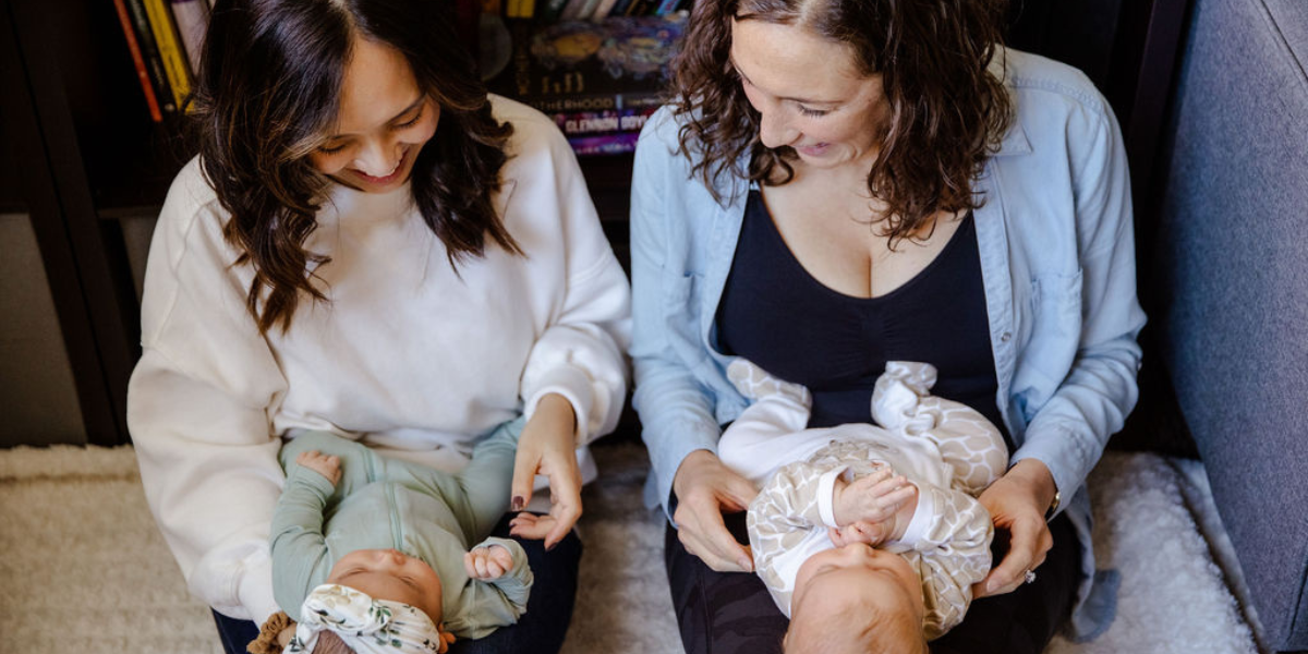 Two new moms sit next to each other holding their newborns.